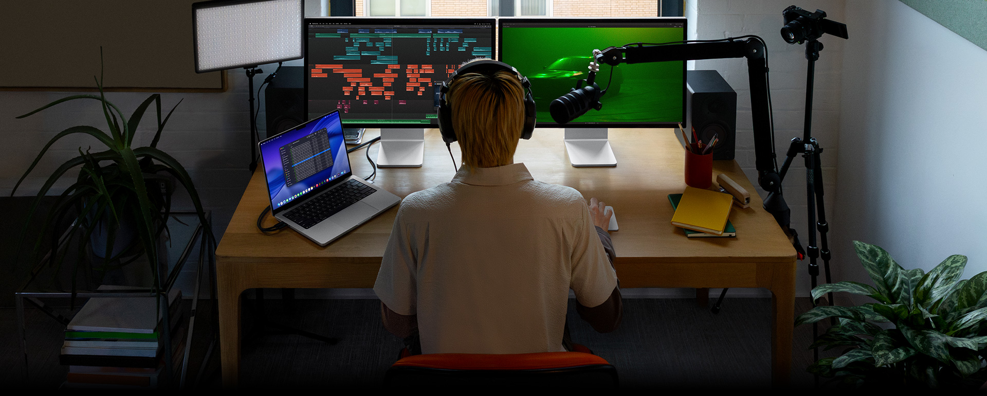 Seseorang sedang duduk di depan meja dengan mikrofon dan headphone di depan tiga perangkat, Mac Studio Display memperlihatkan alat pengeditan audio yang mendetail, klip suara dalam berbagai blok warna, layar Mac Studio Display kedua menampilkan mobil dengan pencahayaan hijau bernuansa sentimental, layar MacBook Pro 16 menampilkan daftar panjang foto dalam proyek Final Cut Pro