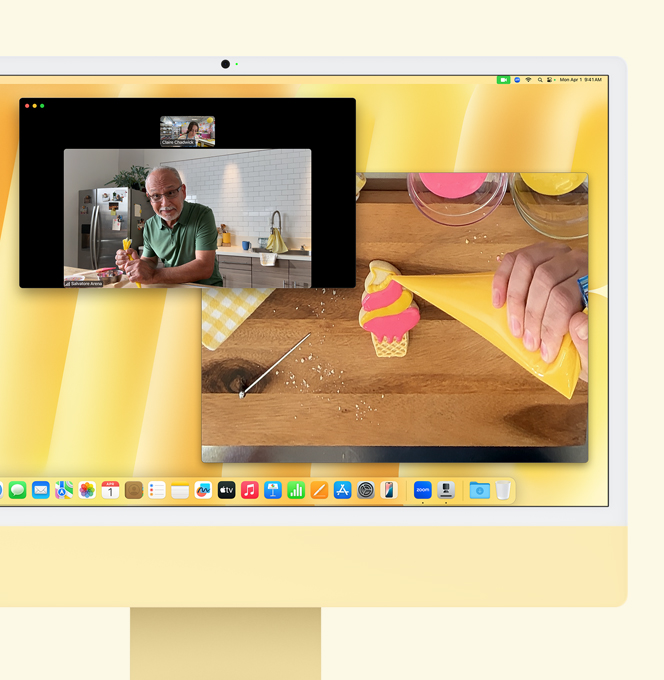 Yellow iMac. Desk View demo. Screen shows baker on video call with another person. One screen displays the baker and the man's face. The other screen displays the baker's hands icing a cookie