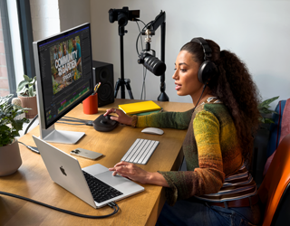 A woman in her home working at her desk with MacBook Pro connected to two external displays, wearing headphones and speaking into a microphone