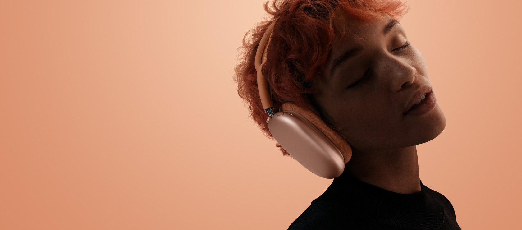 A person with head tilted and eyes closed wearing AirPods Max 2, orange colour, over orange-pink background