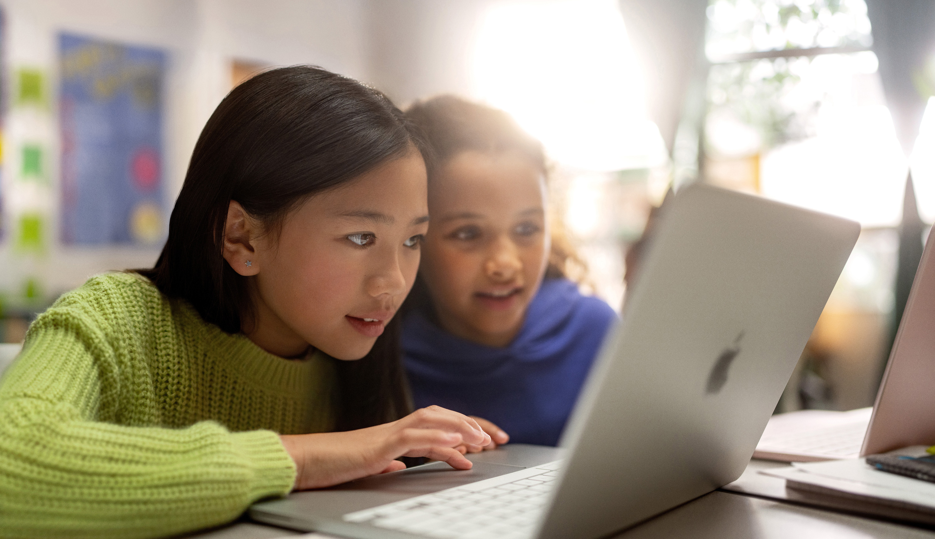 Dos jóvenes estudiantes en un escritorio miran con atención una MacBook mientras trabajan en un salón de clases
