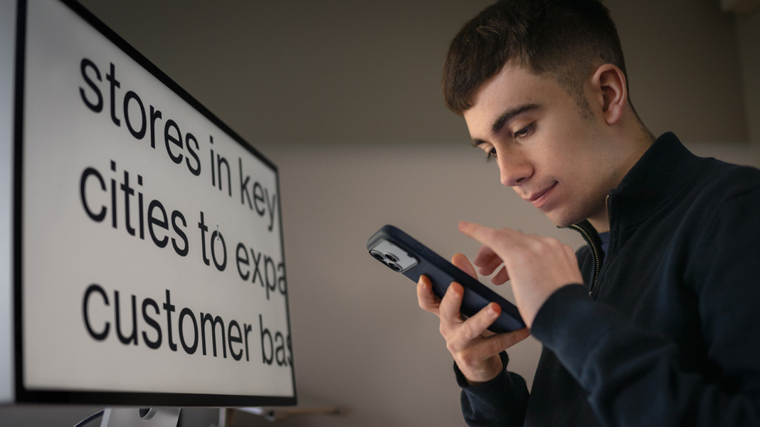 A student uses an iPhone while sitting in front of a monitor displaying content from his business class in large, zoomed‑in text