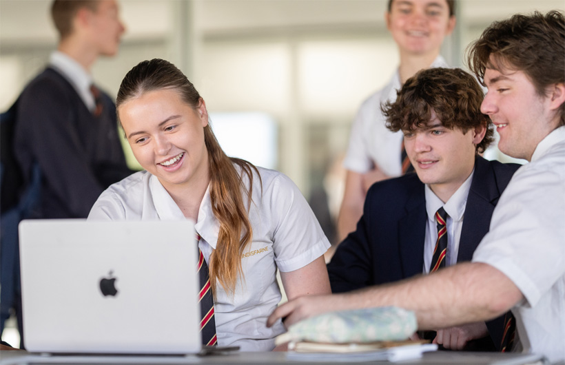 Leerlingen in schooluniformen zitten rond een MacBook en werken lachend samen.