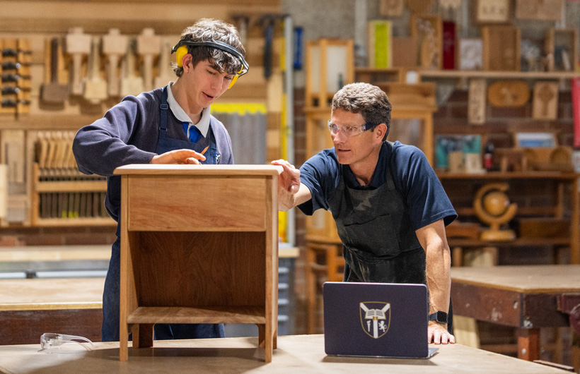 Een houtbewerkinginstructeur en leerling onderzoeken een klein houten kastje. Een schoollaptop staat voor hen op de werkbank.