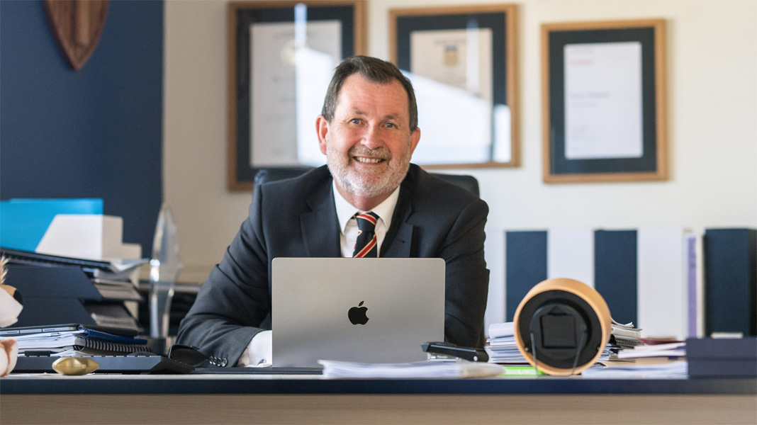 Een directeur zit achter zijn bureau achter een geopende Apple laptop en lacht naar de camera.