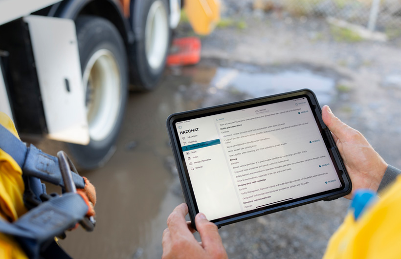 一部雙手握持的 iPad，螢幕上開啟了 HazChat app，顯示 Job Details、Planning、Hazards and Controls 等資訊項目；旁邊有另一名作業人員的身影，背景是現場的泥濘環境