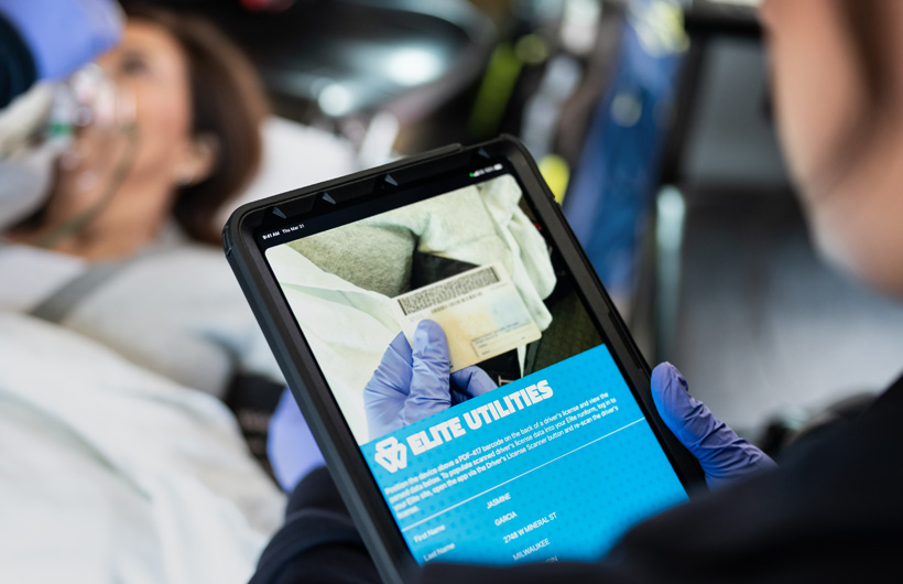 The Elite Utilities app is open on iPad, and a paramedic scans a patient's driving licence, while the patient lays on a transport bed