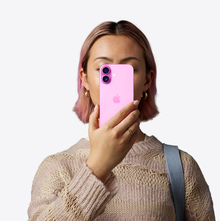 Une personne tenant un iPhone 16 en finition rose face à son visage montrant la face arrière, système avancé à deux caméras dans le coin supérieur gauche.