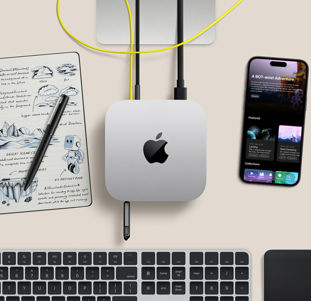 Mac mini, top, silver aluminum, rounded corners, glossy black Apple logo centered, connected by cord, USB stick inserted in USB‑C port in front, Studio Display stand partially visible, iPhone, Magic Trackpad, Magic Keyboard with Touch ID, stylus and e-ink sketch pad
