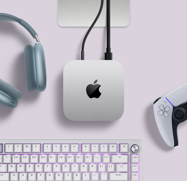Mac mini, top, silver aluminium, rounded corners, glossy black Apple logo centred, connected by cord, Studio Display stand partially visible, AirPods Max 2, blue colour, PlayStation DualSense controller, white color, and white keyboard