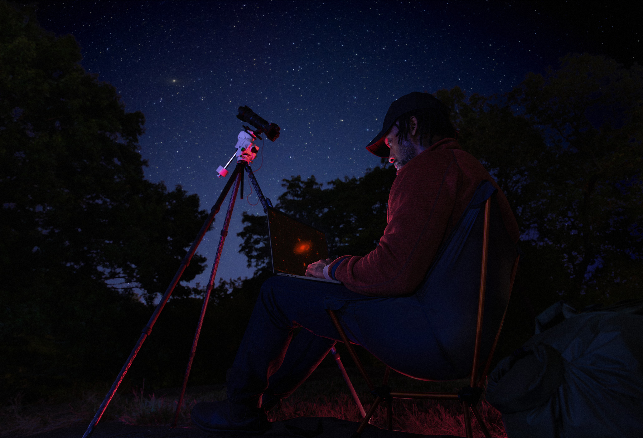 Una persona que está al aire libre, de noche, tiene un MacBook Pro apoyado en su regazo conectado a un dispositivo con un trípode que apunta hacia las estrellas