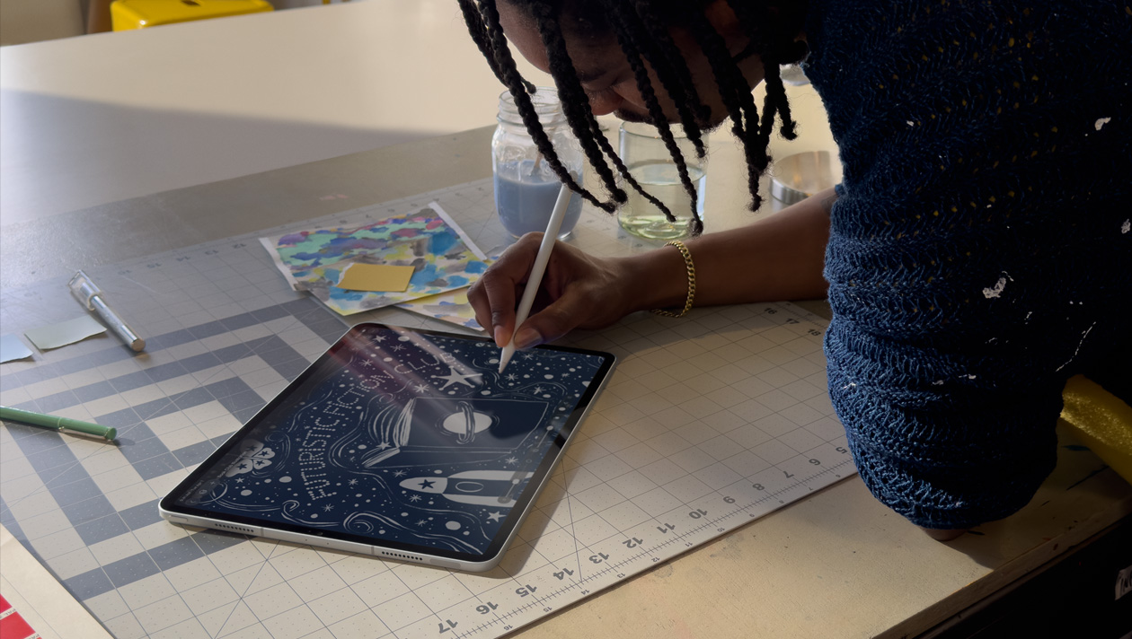 Person with braids wearing a blue sweater drawing on iPad Pro with Apple Pencil, screen shows illustration of a night sky with a space ship, a butterfly, a planet on a book cover, and futuristic fiction club written in the centre